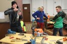 3 Personen in einer Werkstatt, auf dem Tisch liegt eine Motorsäge und Werkzeug